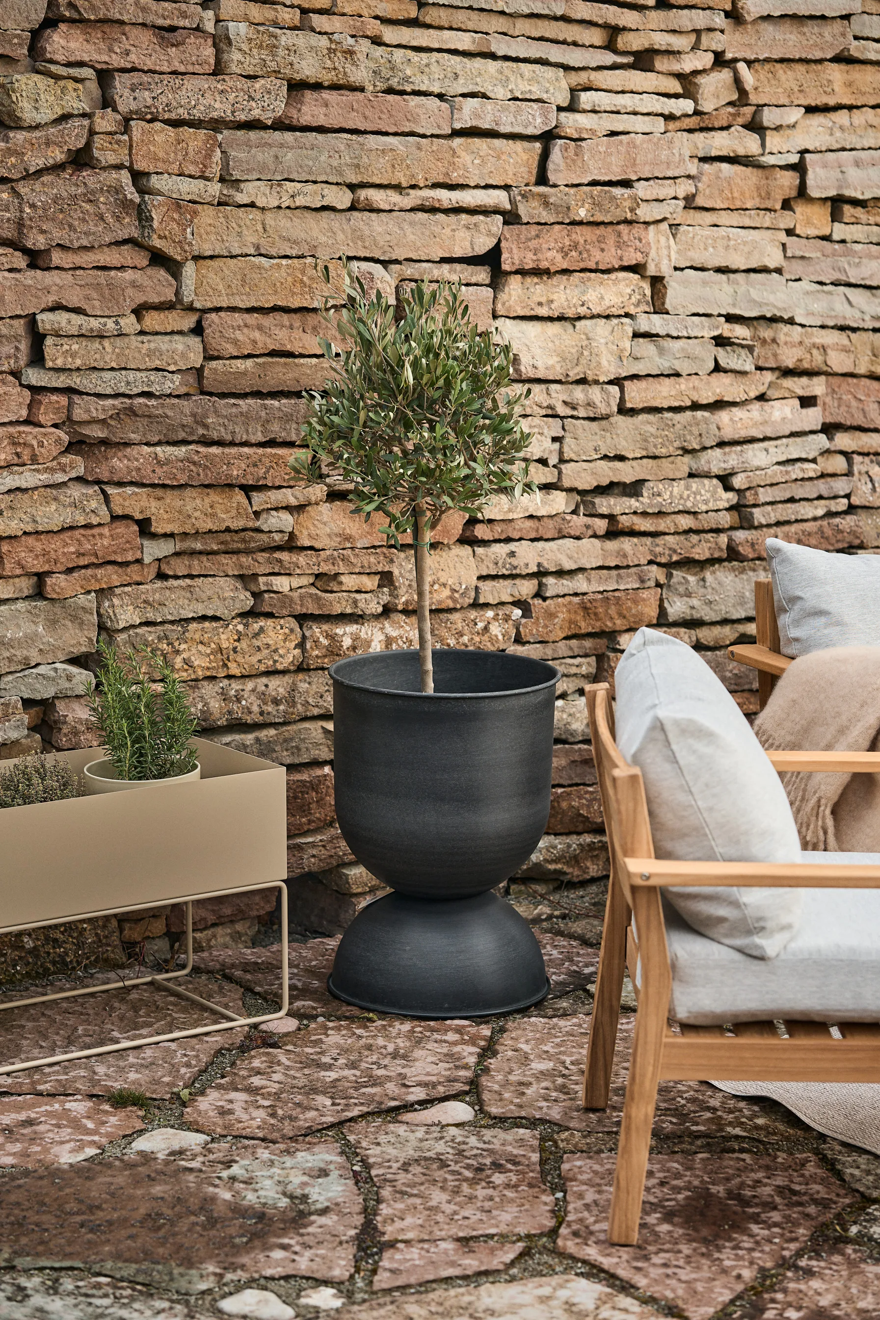 Small tree in a black hourglass planter and a beige rectangular planter box against a rustic stone wall.
