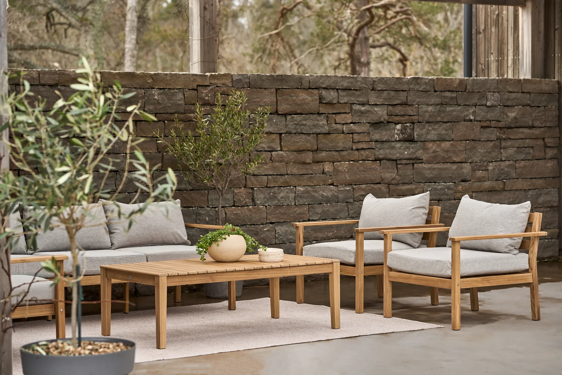 Modern wooden patio furniture set with gray cushions and a coffee table in front of a stone wall.