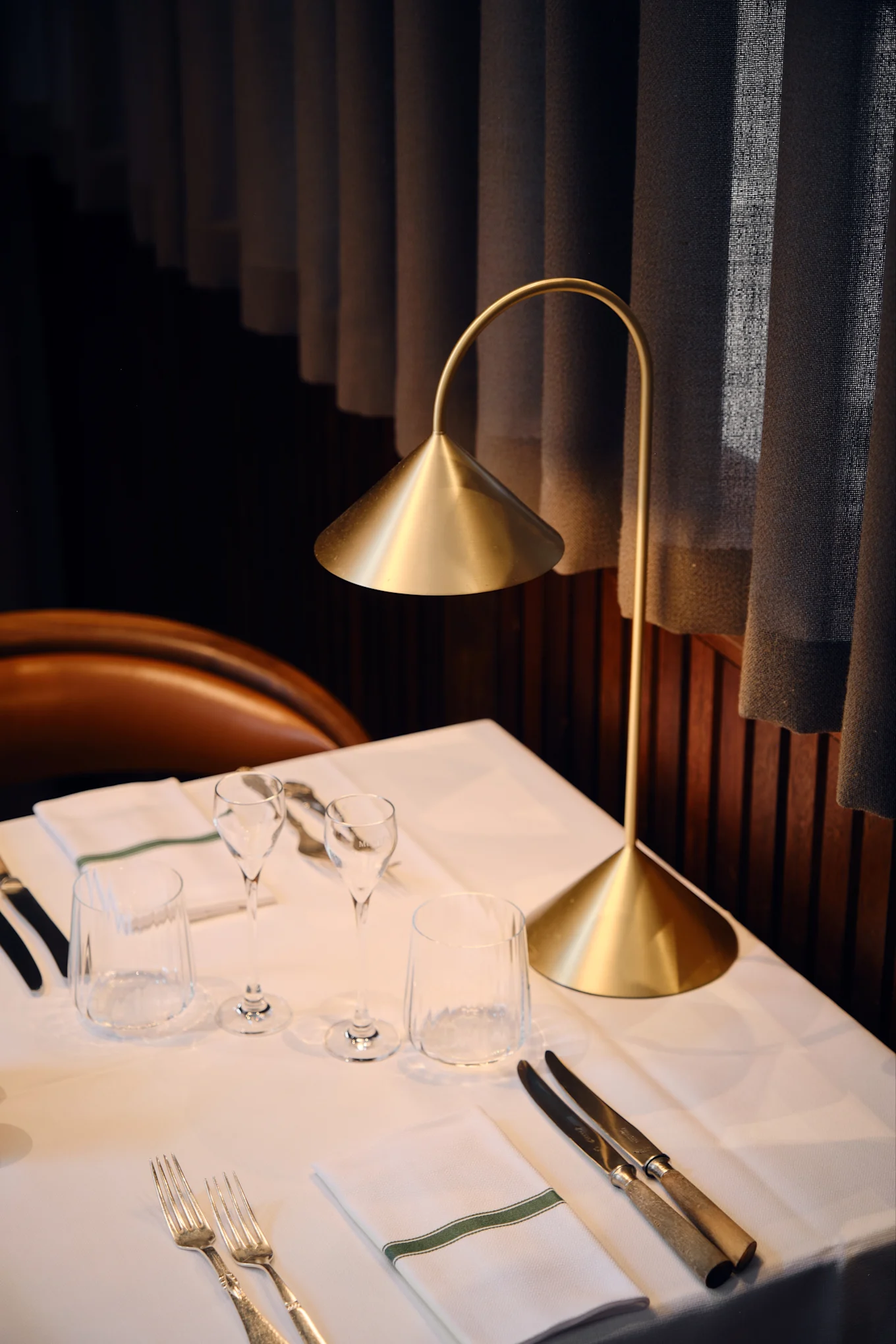 A modern gold table lamp illuminates a set restaurant table with white linens, glasses, silverware, and a napkin with a green stripe.