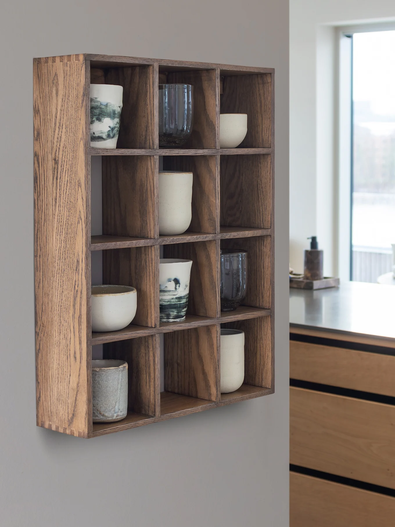 Wooden cubby wall shelf with 12 compartments holding ceramic cups and mugs. A modern kitchen and window are in the background.