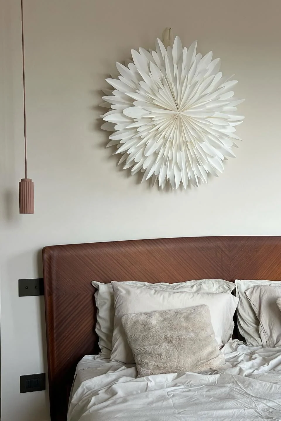 Bedroom with a bed, wooden headboard, white bedding, fluffy pillows, two pendant lights, and a white paper star wall decoration.