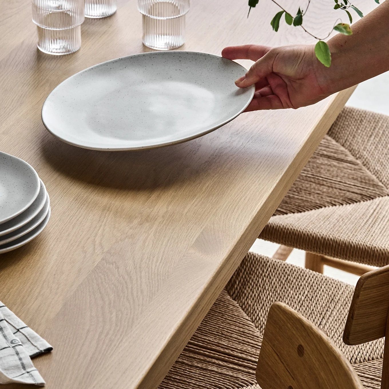 A hand places a speckled grey oval plate on a light wooden table, with stacked bowls and ribbed glasses nearby. A woven chair is to the right.