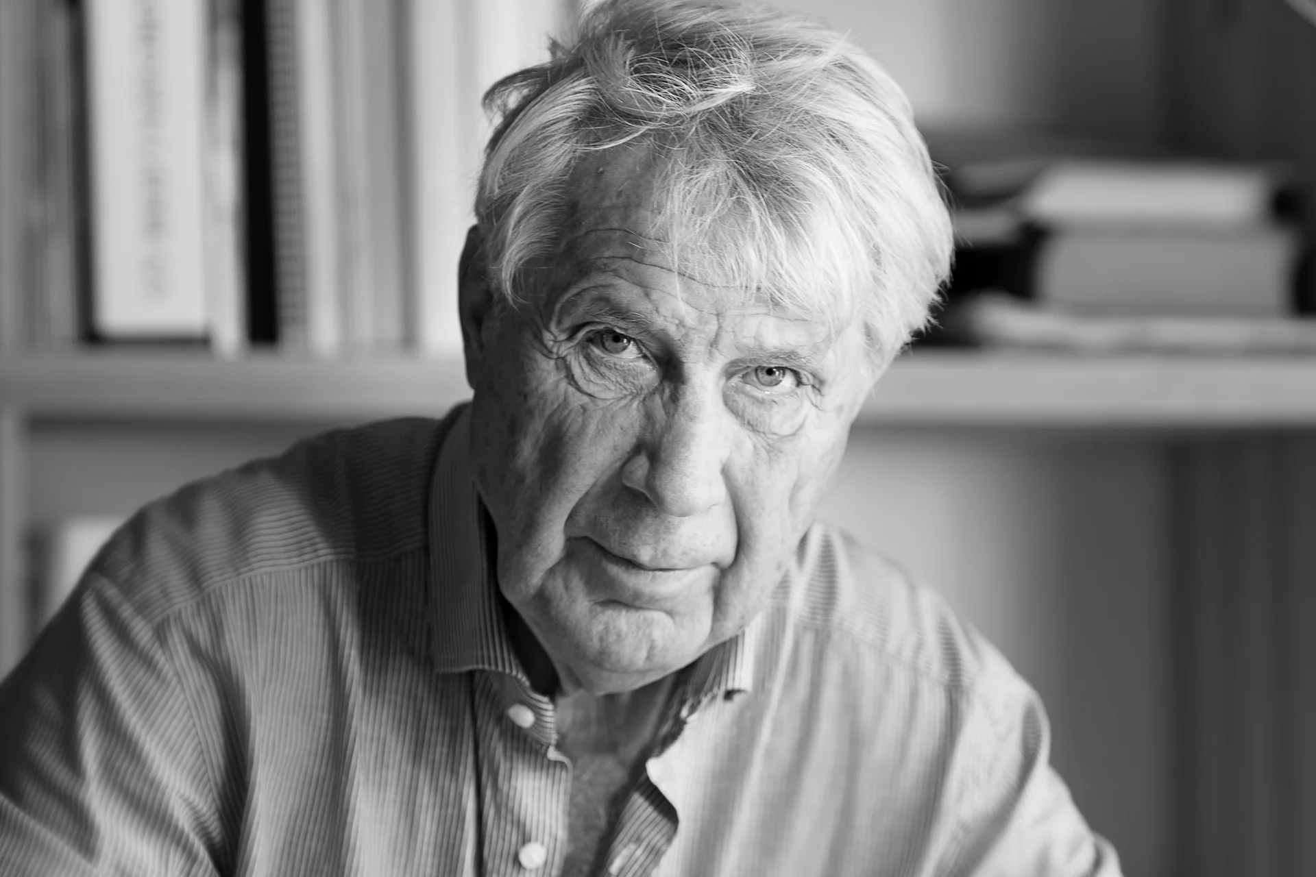 Black and white close-up of an older man with gray hair in a striped shirt, looking intently at the camera.