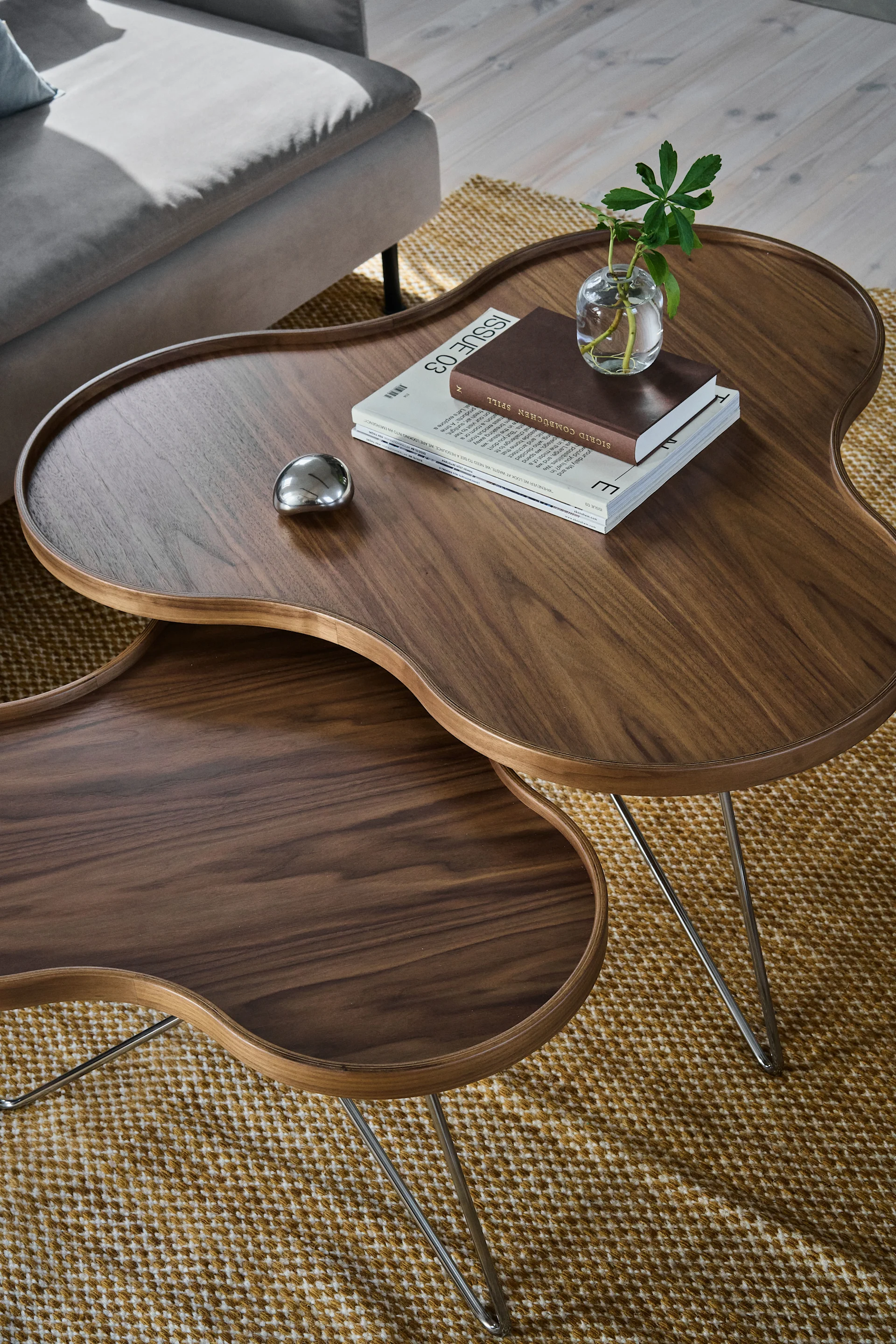 Two dark wood, kidney-bean-shaped nesting coffee tables on a patterned rug, with a plant, books, and a metallic object.