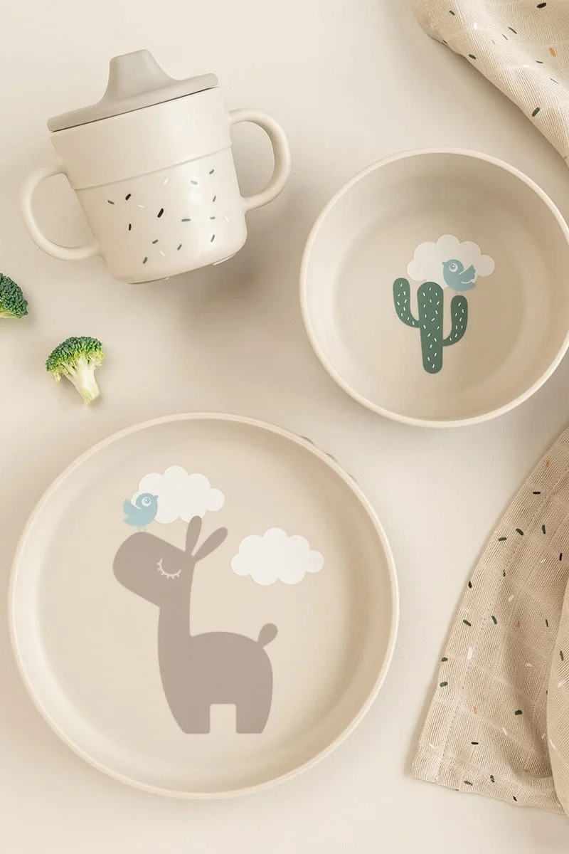 A light beige children's dinnerware set with a sippy cup, a bowl with a cactus, and a plate with a llama and birds.