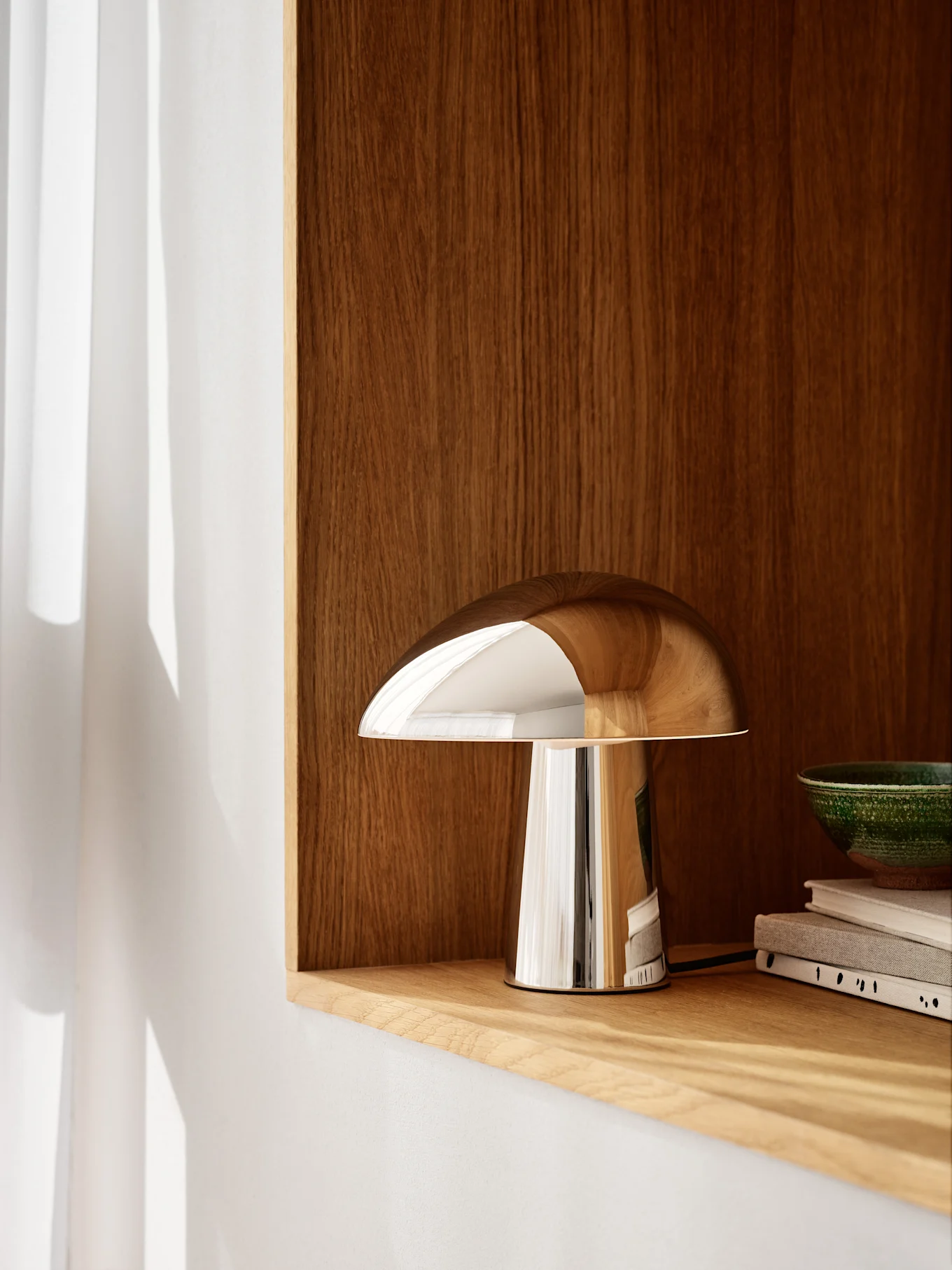 A polished chrome mushroom-shaped lamp sits on a light wooden shelf, with a dark wooden wall behind it. A green bowl and books are to the right.