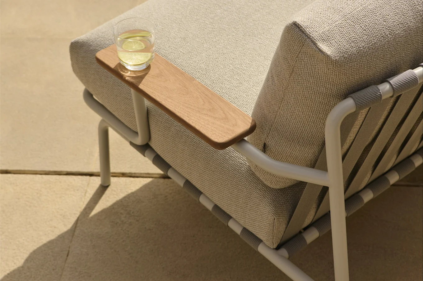 Outdoor lounge chair with light beige cushions, a light metal frame, and a wooden armrest holding a glass of water with lime.