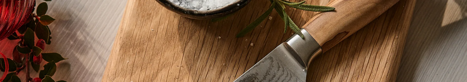 Knife, rosemary, and sea salt on a wooden board next to a red festive decoration.