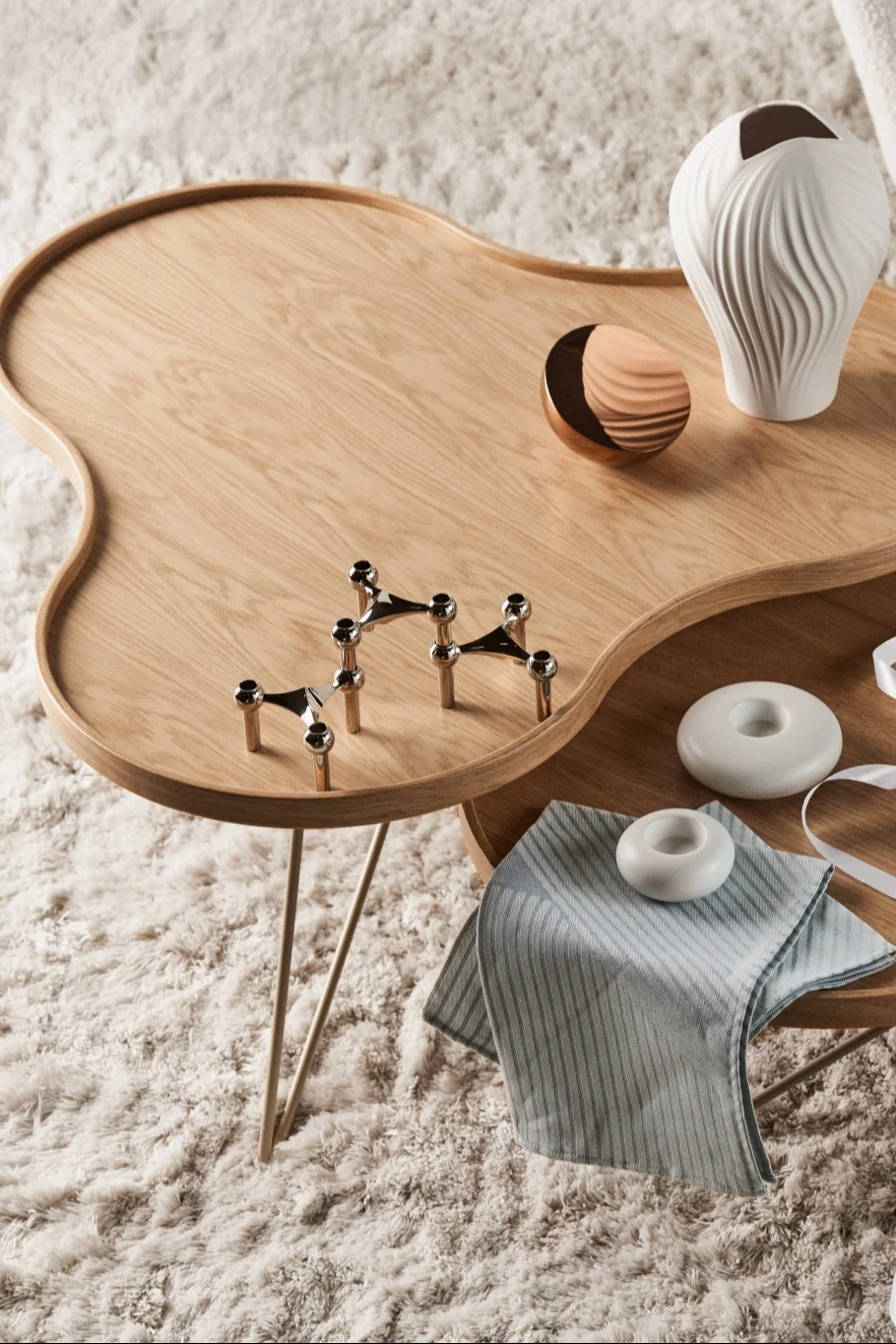 An organically shaped wooden coffee table on a fluffy rug, decorated with a white vase, copper bowl, and silver candle holders.