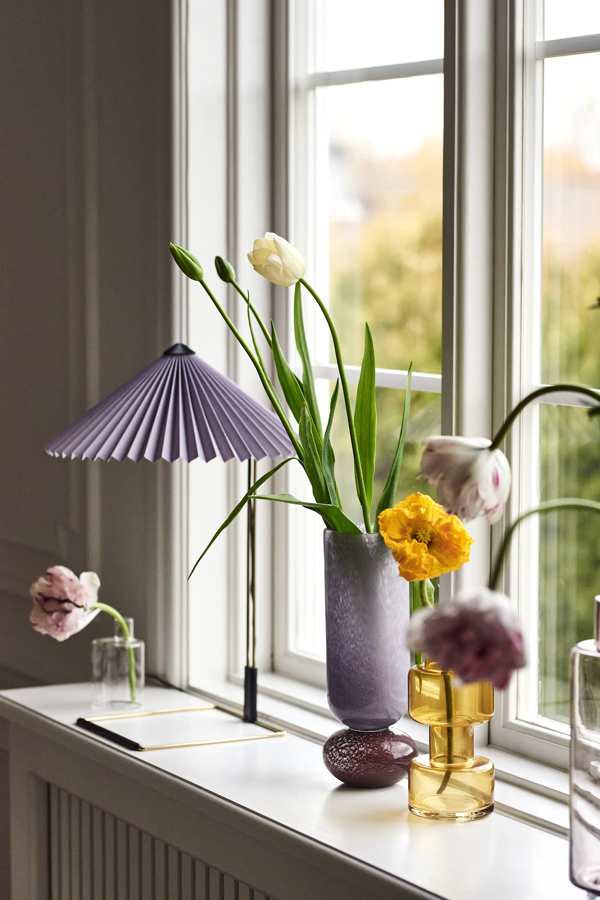 A collection of vases with various flowers on a window sill, including white tulips, yellow poppies, and pink tulips, next to a purple pleated lamp.