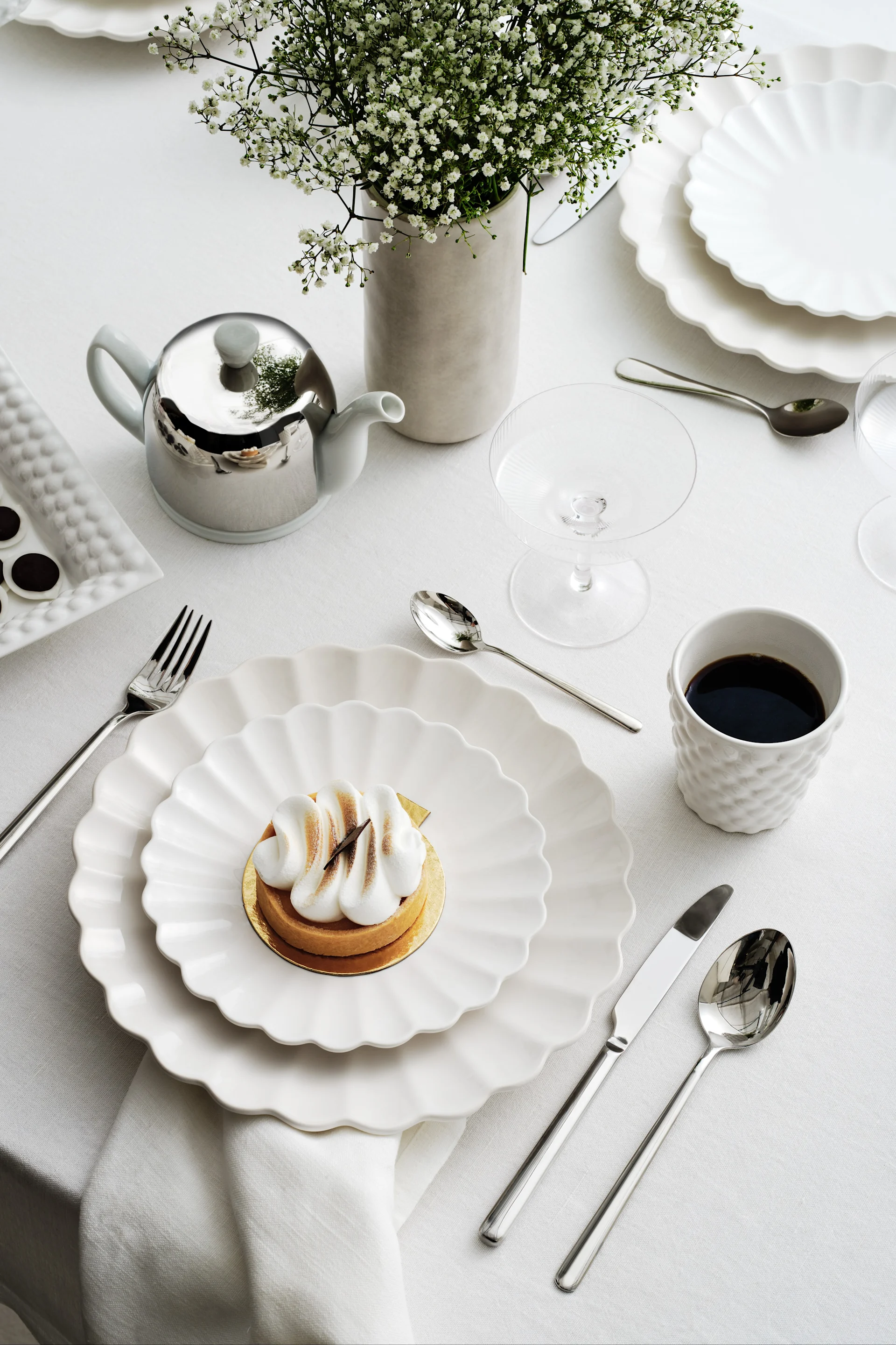 Elegant table setting with white scalloped plates, a meringue tart, coffee, a silver teapot, and baby's breath flowers.