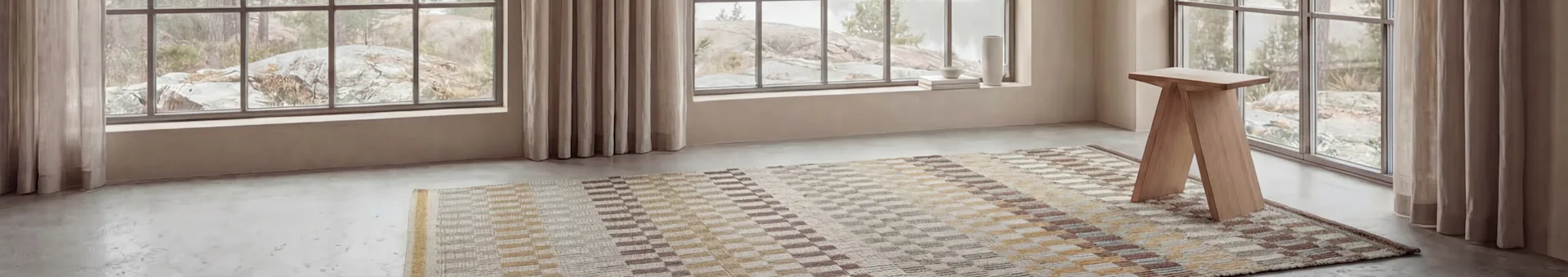A room with large windows revealing a rocky landscape. A patterned rug and a light wooden stool on a grey floor, with decor on the windowsill.