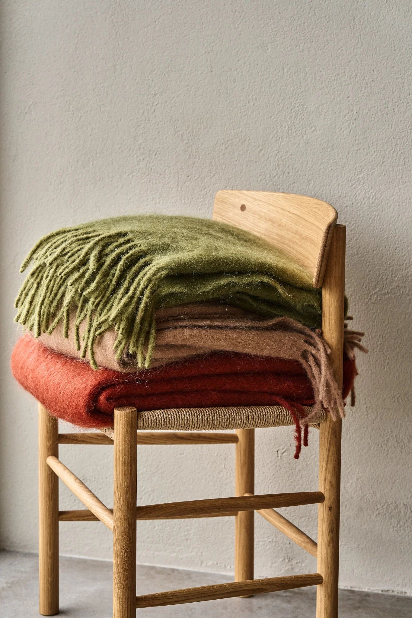Stack of three soft blankets in green, pink, and orange on a light wooden chair with a woven seat.