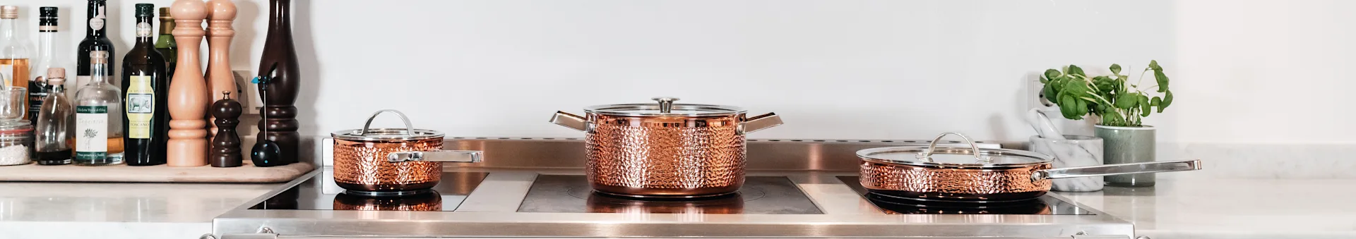 Three hammered copper pots and pans with silver handles sit on a modern stovetop.