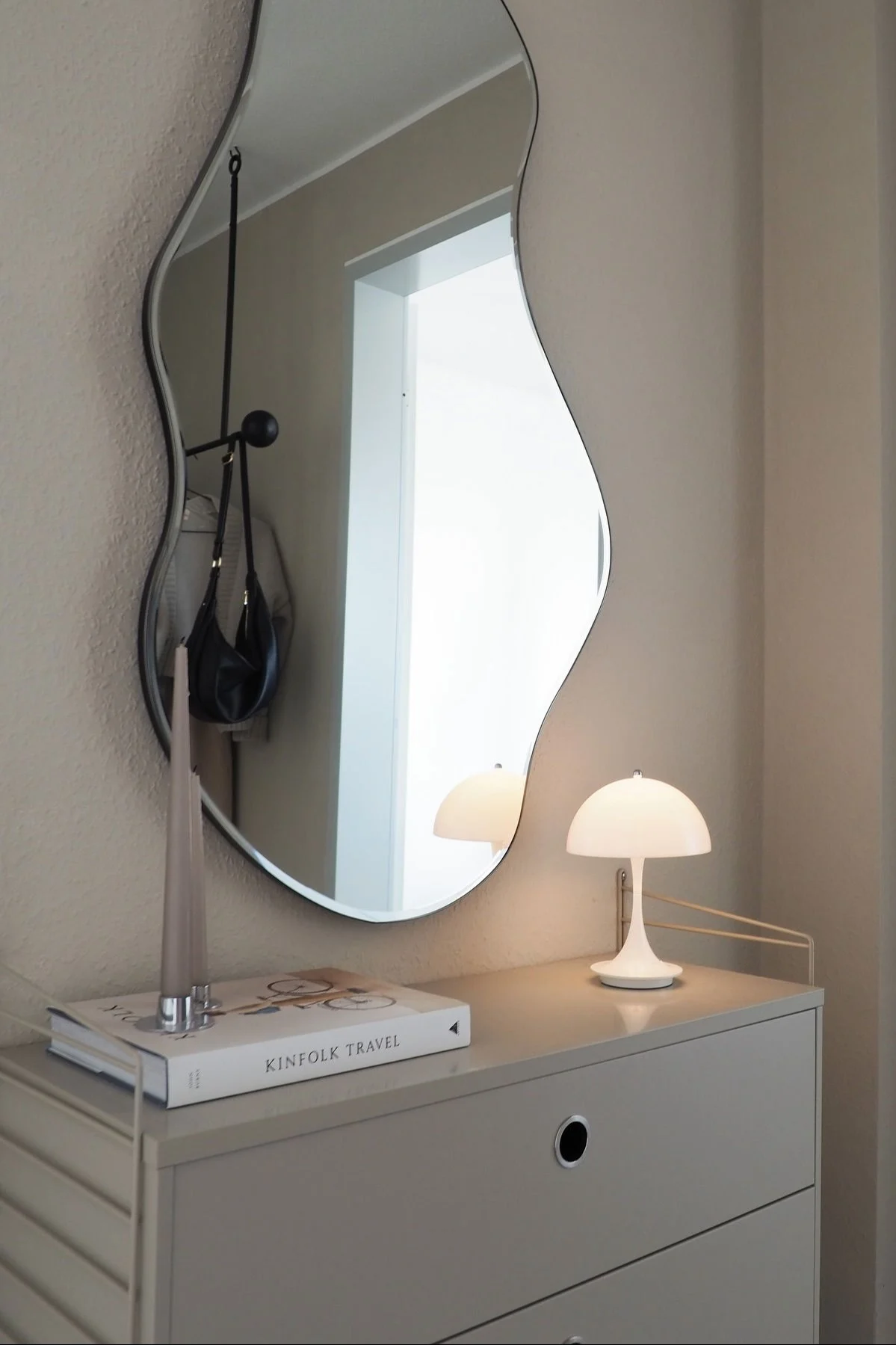 Wavy mirror reflecting a handbag, above a dresser with a mushroom lamp, book, and candle.