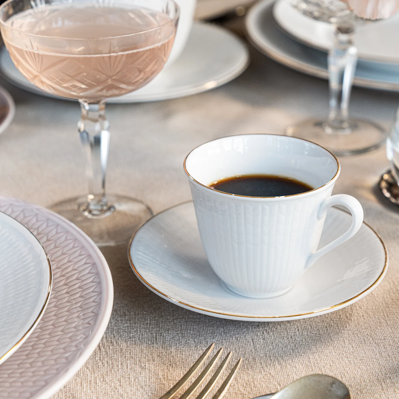 A white coffee cup with a gold rim on a saucer, next to a pink patterned plate and a coupe glass with rosé on a linen tablecloth.