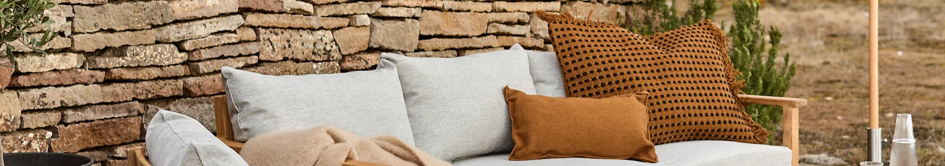 Outdoor sofa with light grey cushions, a brown blanket, and two rust-colored cushions, one with black dots, against a stone wall.