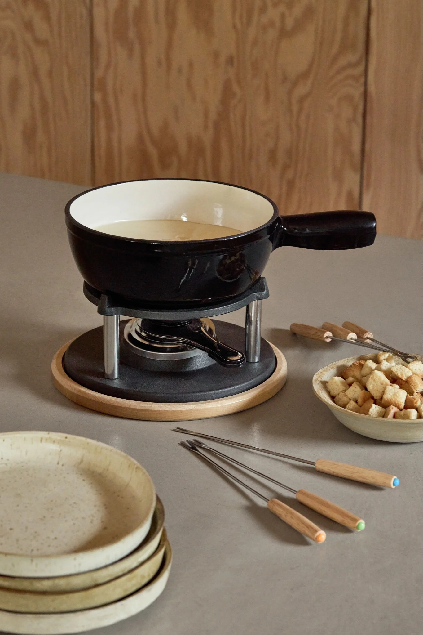 Black fondue set with a pot of melted cheese on a wooden base, surrounded by bread cubes and fondue forks.