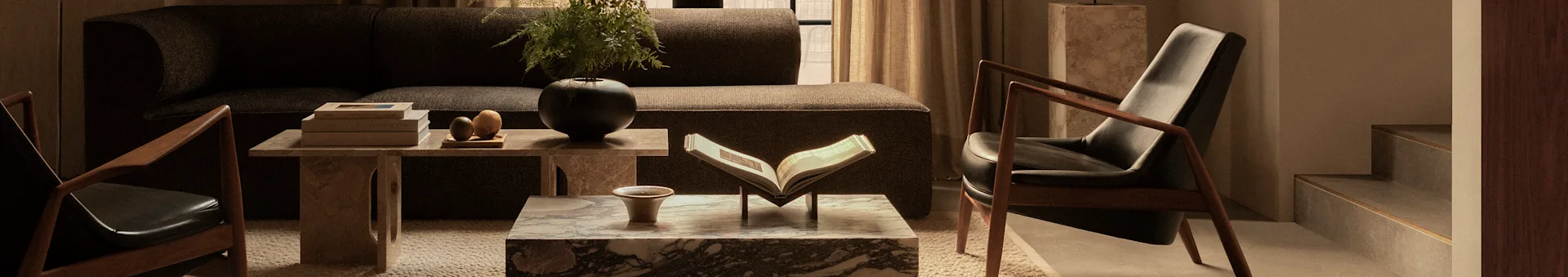 A cozy living room featuring a dark brown sofa, two dark leather armchairs with wooden frames, and marble coffee tables adorned with books and plants.