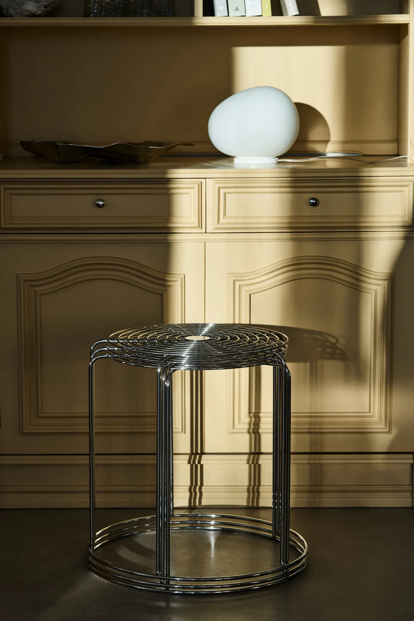 A stack of chrome-plated metal stools in front of a light brown cabinet with a white egg-shaped lamp, bathed in sunlight.