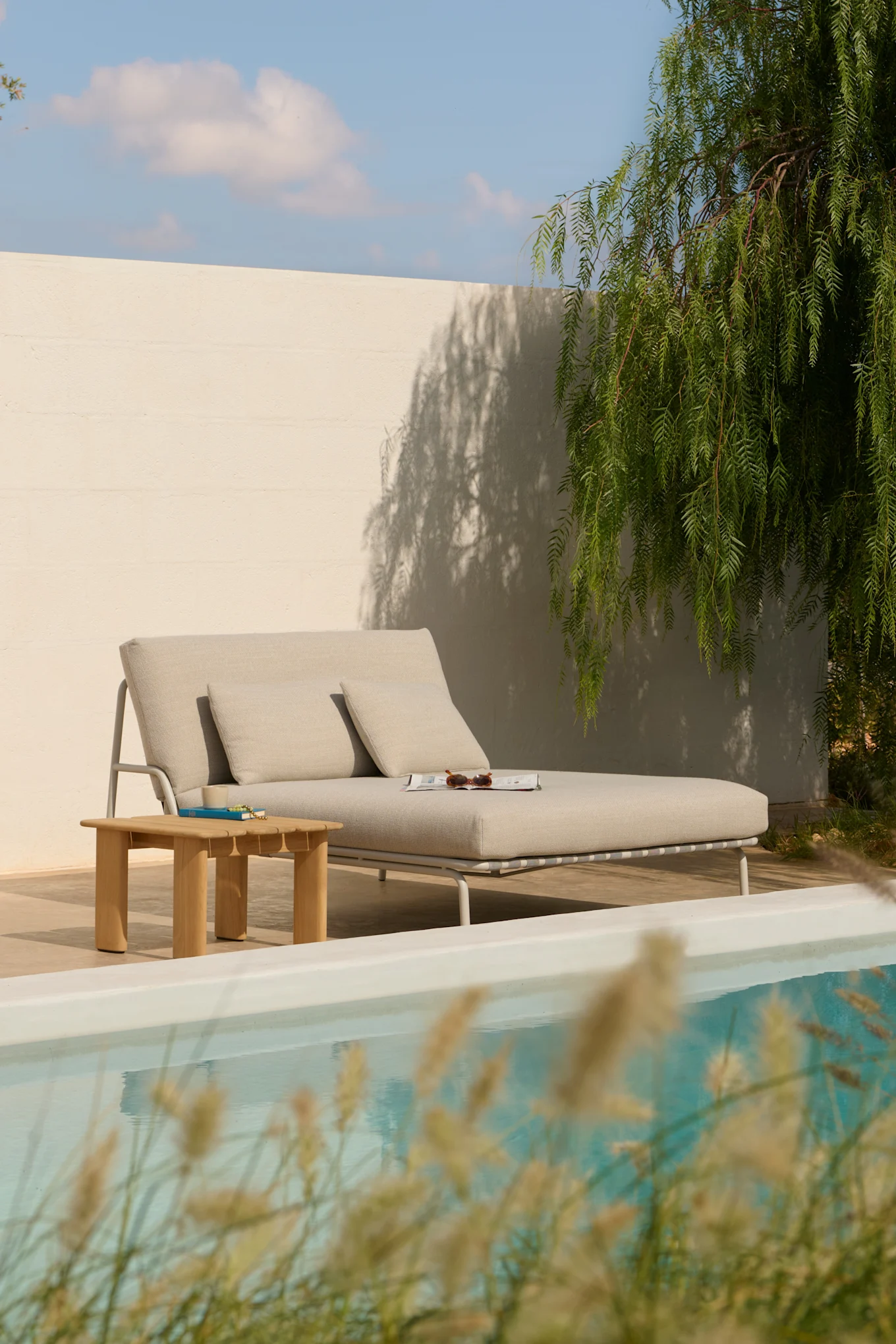 Light gray outdoor chaise lounge with pillows, a wooden side table, and a magazine by a pool.