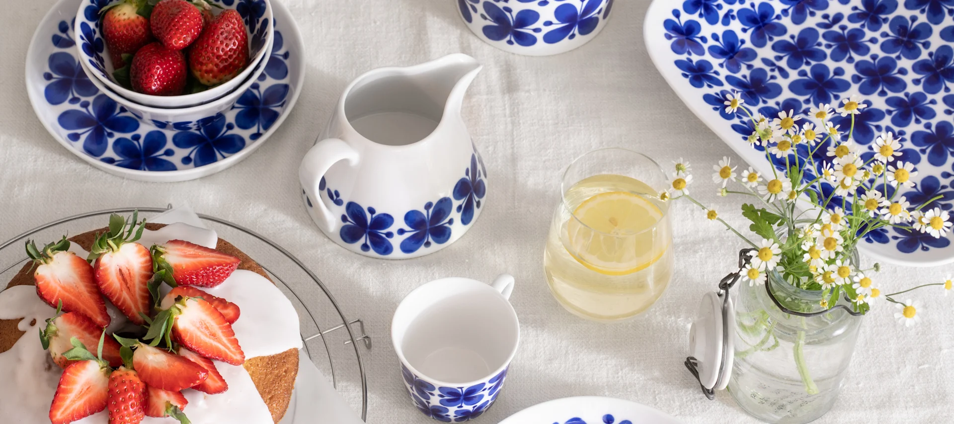 Table with white and blue patterned ceramic dishes, a strawberry cake, a bowl of strawberries, lemon water, and daisies.
