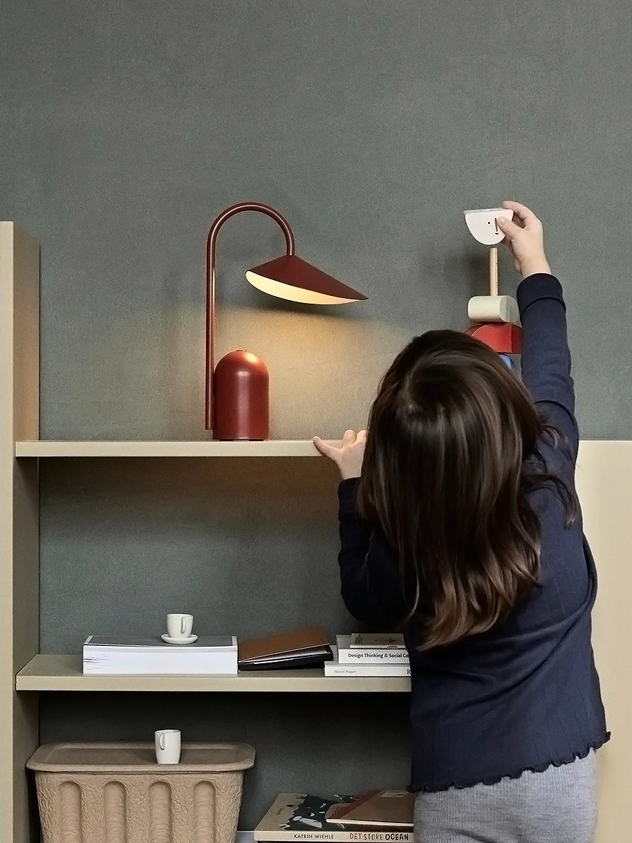 A child reaches for a wooden toy bird on a light shelf with a red lamp and books; a woven basket sits on the floor.
