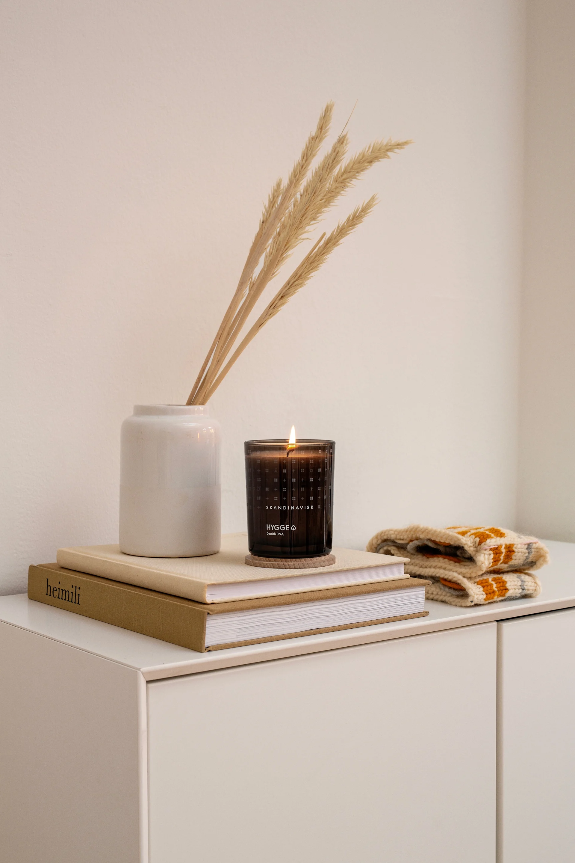 A lit 'Hygge' candle and dried grass in a white vase next to patterned knit fabric on stacked books.