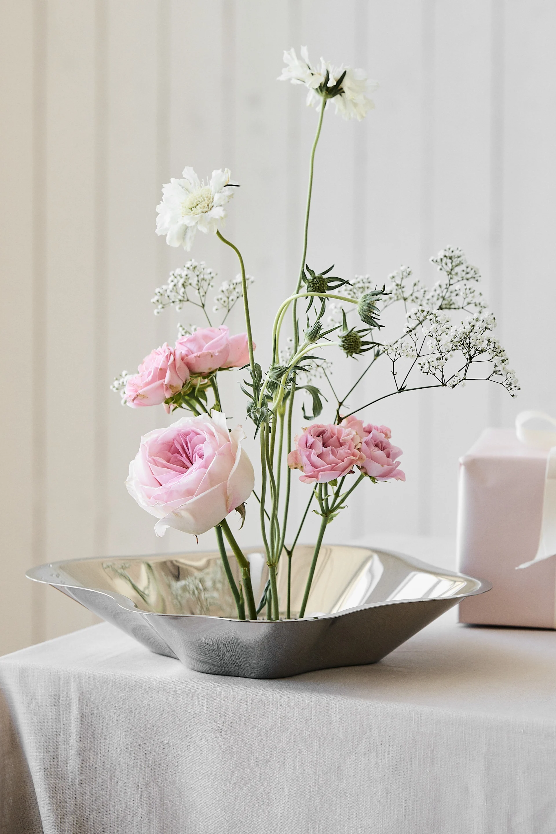 Pink roses and white flowers arranged in a modern silver bowl, with a pink gift box and white ribbon in the background.