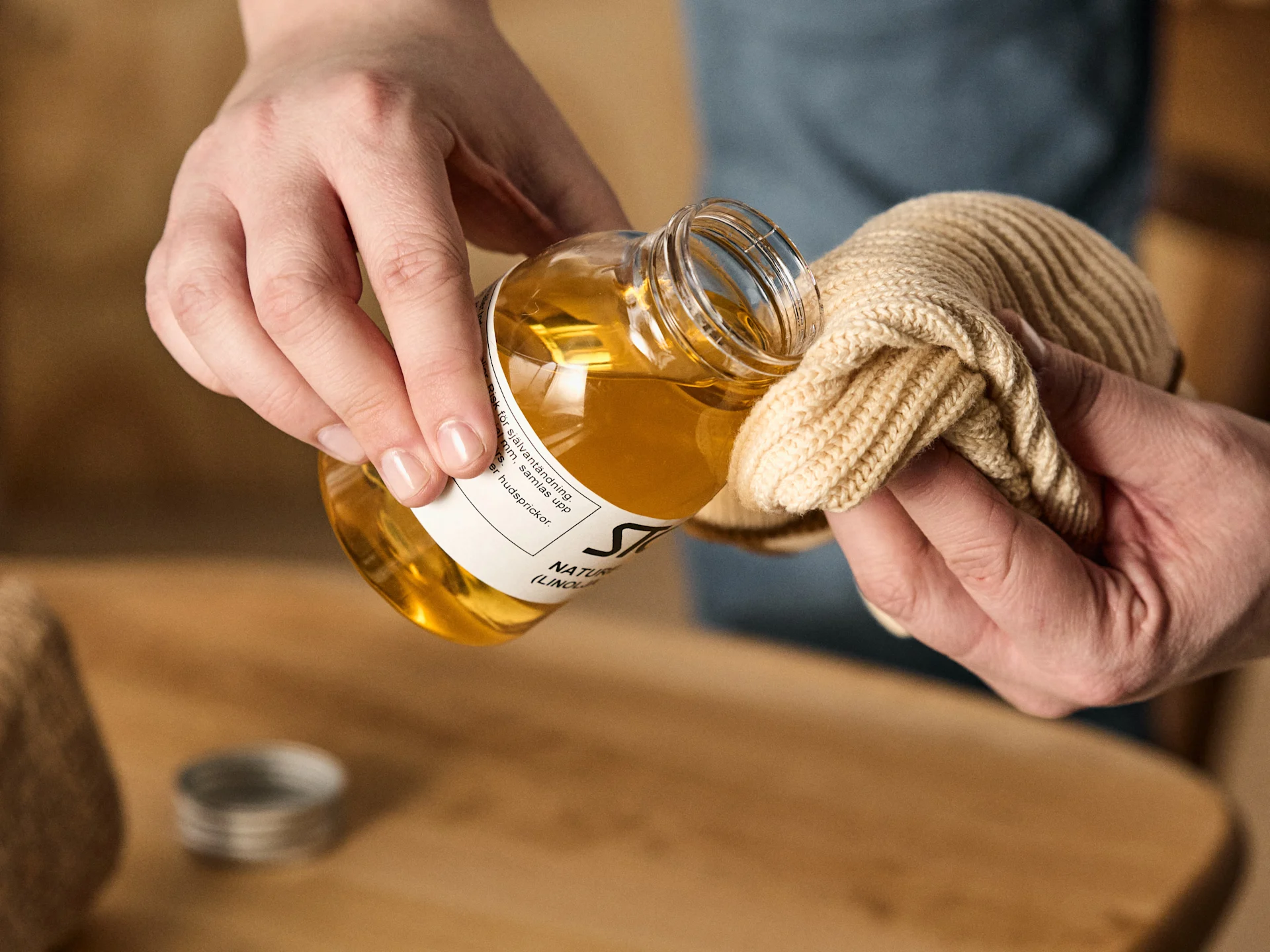 Hands pour golden oil from a clear bottle onto a beige knitted cloth.