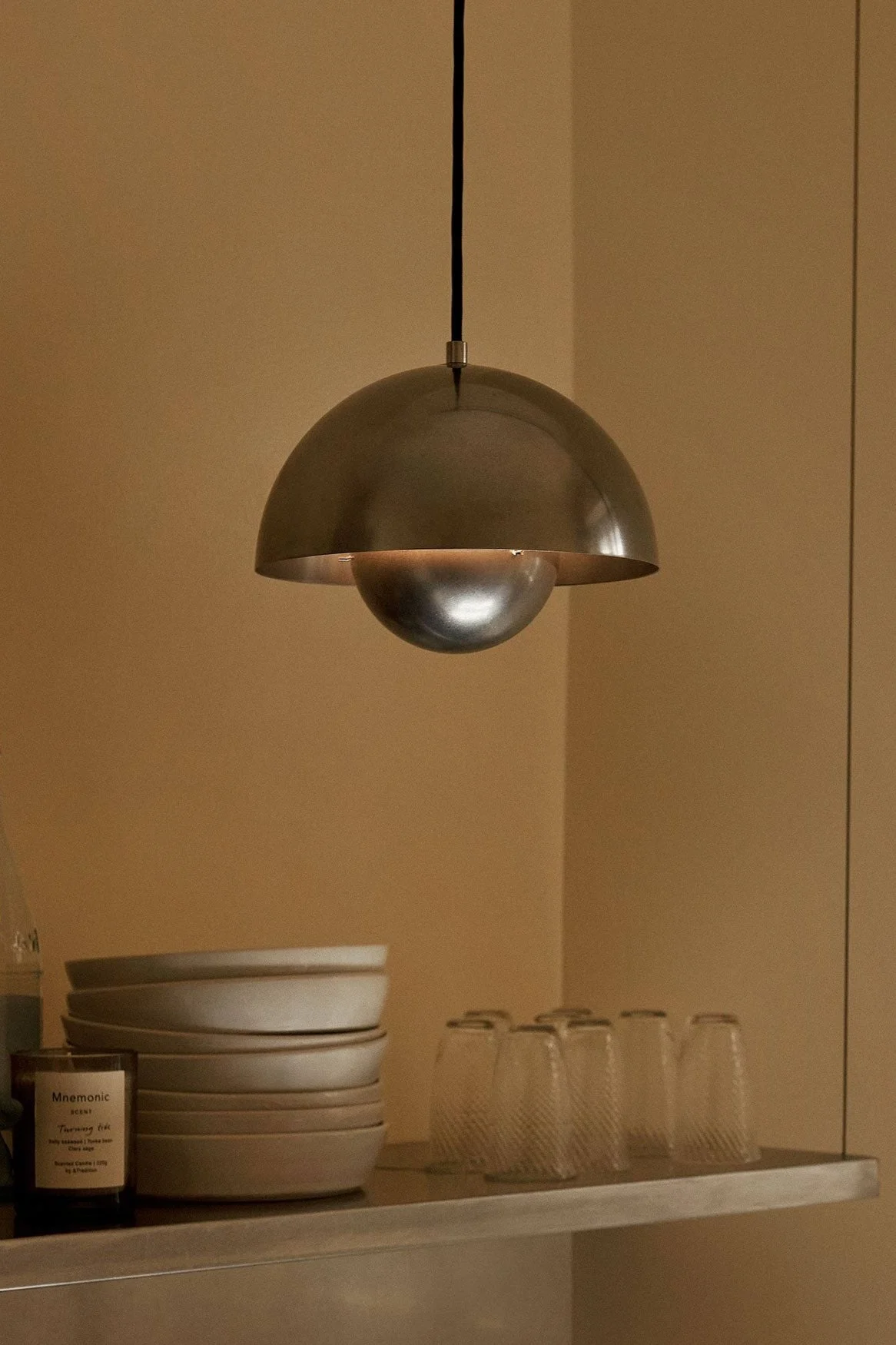 A dome-shaped metal pendant light hangs over a shelf with bowls, glasses, bottles, and a candle on a warm brown wall.