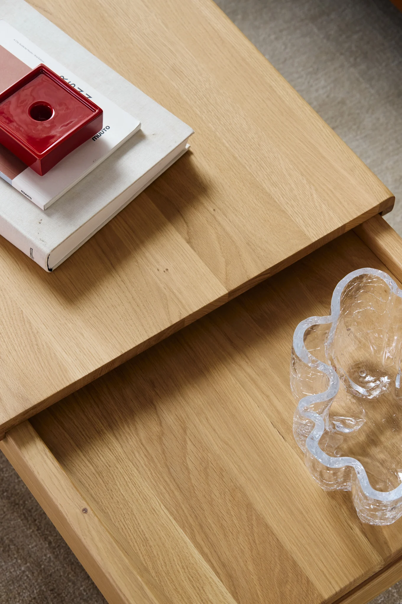 High-angle view of a light wooden table with a red square holder on books and a wavy clear glass bowl in an open drawer.