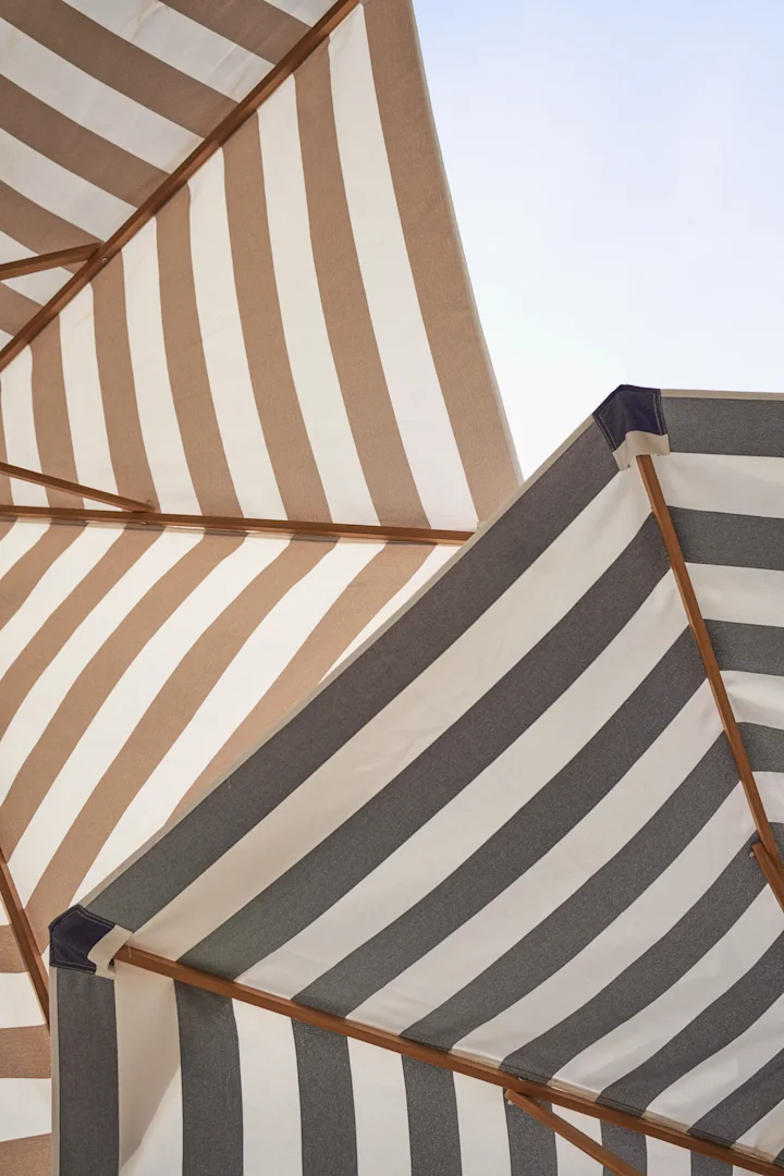 Two overlapping striped beach umbrellas, one brown and white, the other dark grey and white, with wooden frames against a light sky.