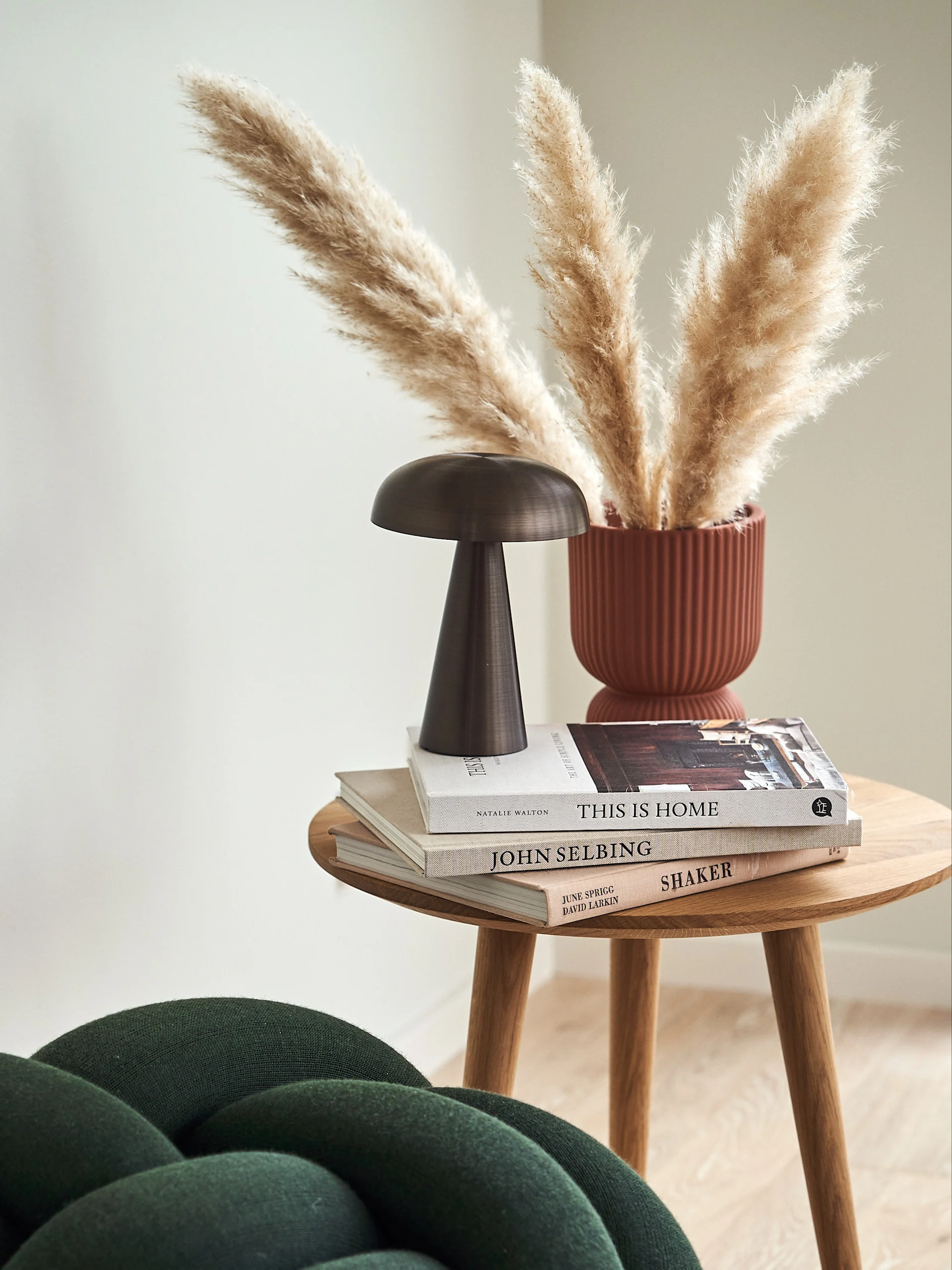 Round wooden side table with stacked books, a dark bronze mushroom lamp, and a rust-colored vase holding pampas grass.