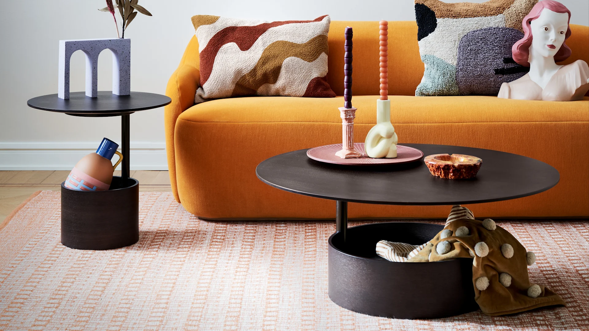 A vibrant living room with an orange sofa, dark side tables, a grey arched vase, colorful candles, patterned pillows, and a textured rug.