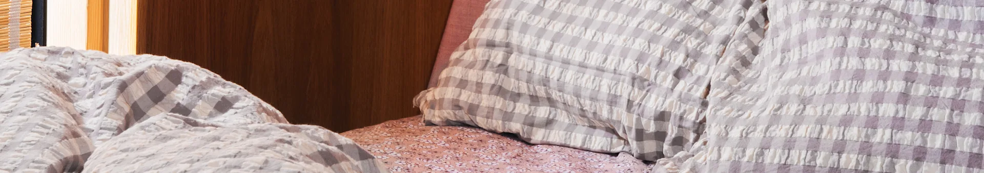 Close-up of a bed with light purple and white checkered comforter and pillows, over a pink floral sheet.