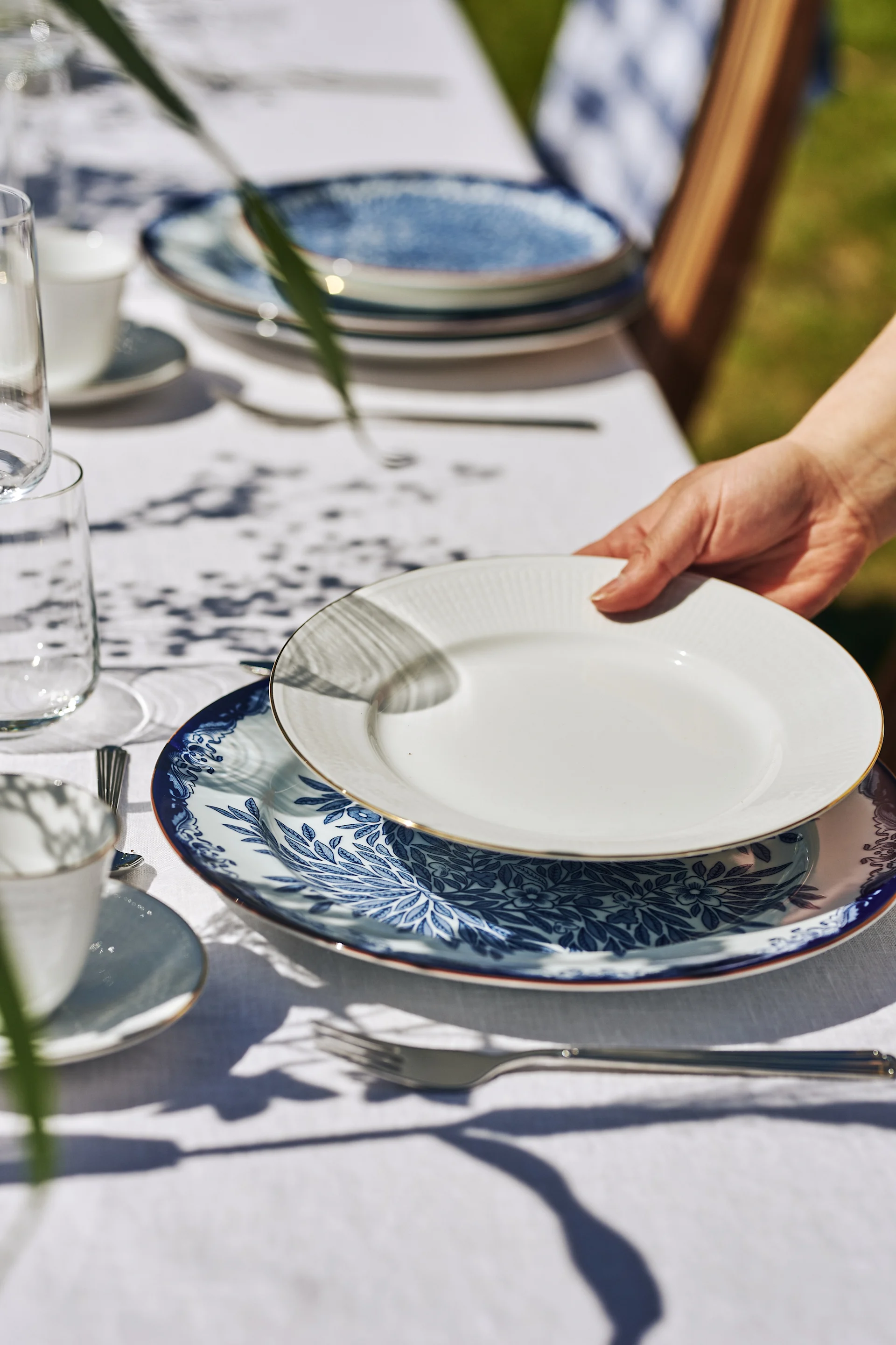A hand places a white plate with a gold rim on a blue and white patterned plate, set on a white tablecloth outdoors.
