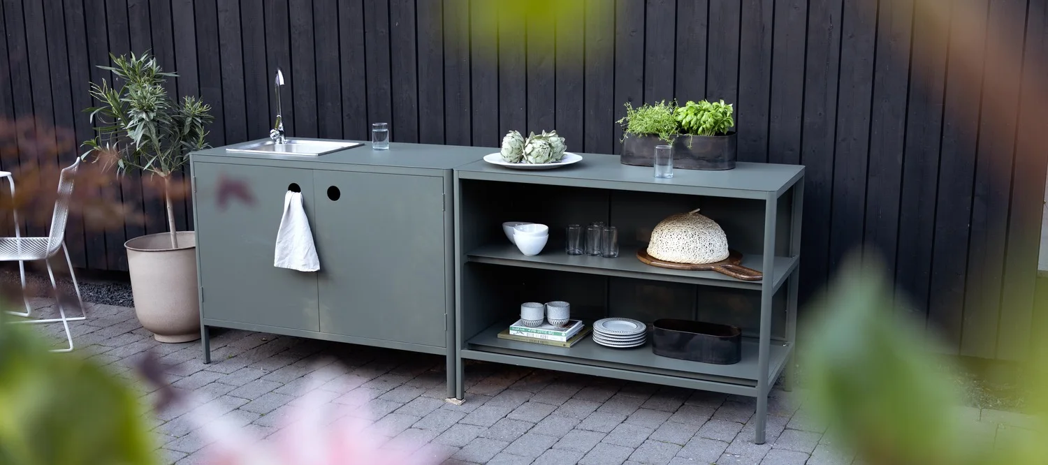 An outdoor kitchen unit in a grey-green color, with a sink and a towel on the left, and shelves displaying kitchenware and potted herbs on the right.