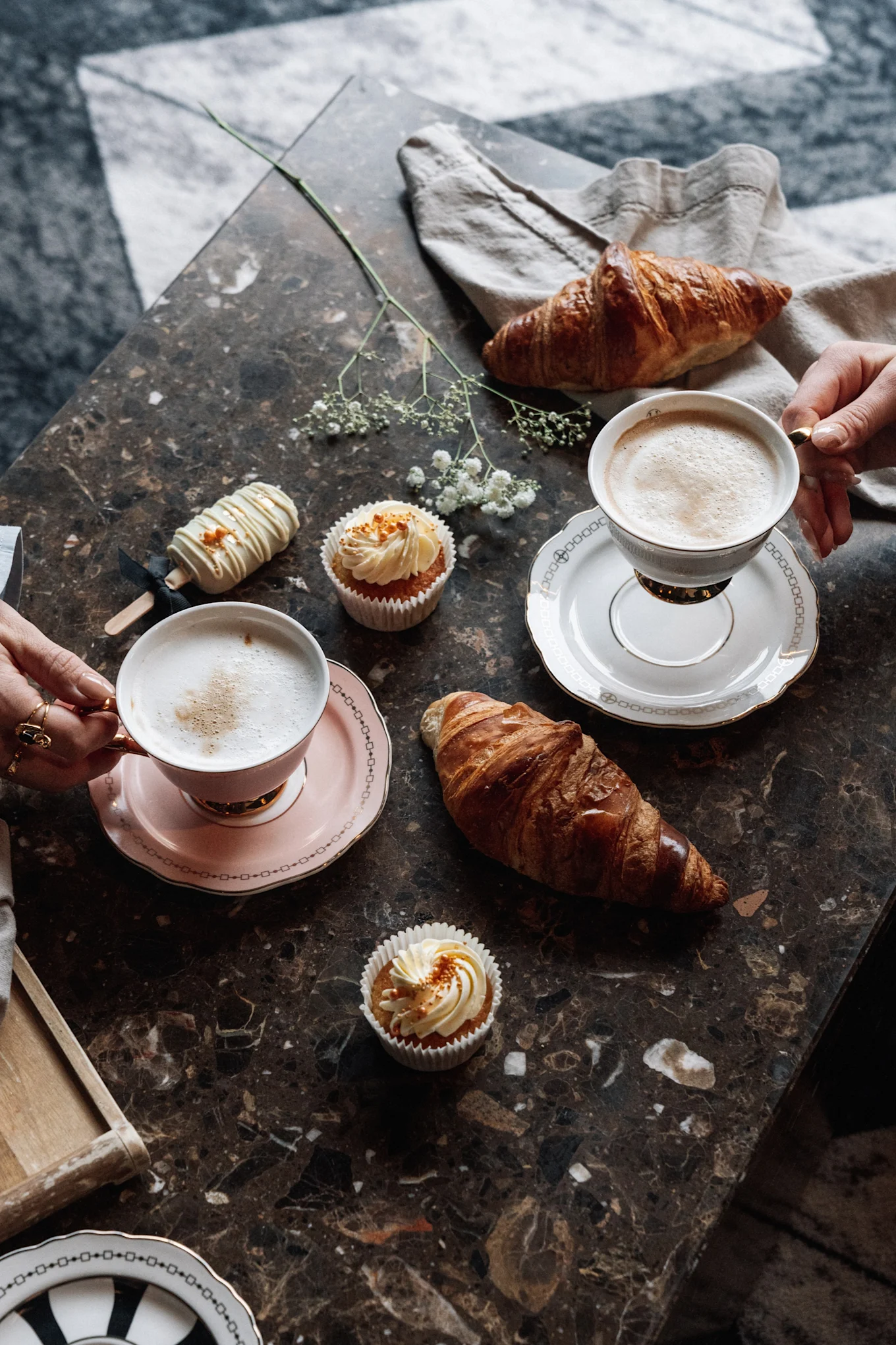 Hands holding coffee cups with croissants, cupcakes, and a cake pop on a marble table.