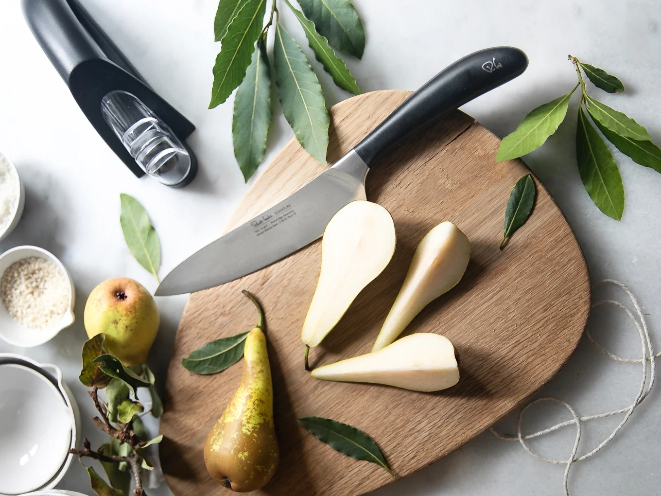 Knife, whole and sliced pears on a wooden cutting board. Bay leaves, a wine pourer, and ingredient bowls surround it.