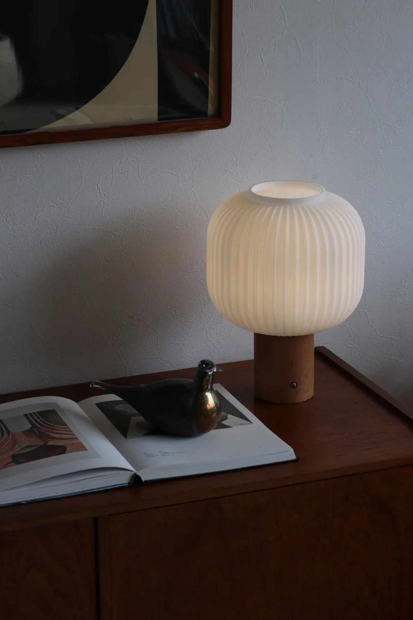 A glowing white ribbed table lamp with a wooden base on a dark cabinet next to an open book and a bird figurine.
