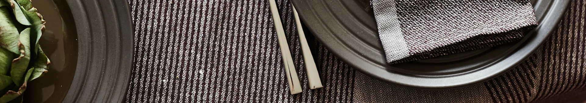 Dining setting with an artichoke on a dark plate, striped placemat, cutlery, and a dark plate with a patterned napkin.