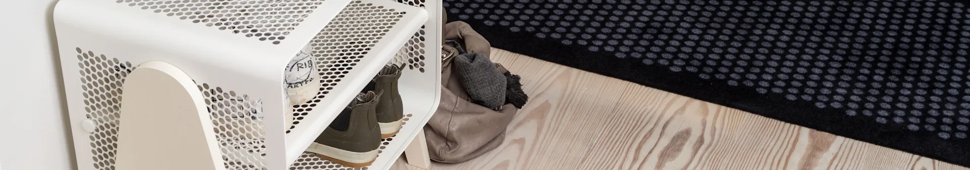 White perforated shoe rack with two pairs of shoes and a beige handbag on a light wooden floor with a black and grey dotted rug.