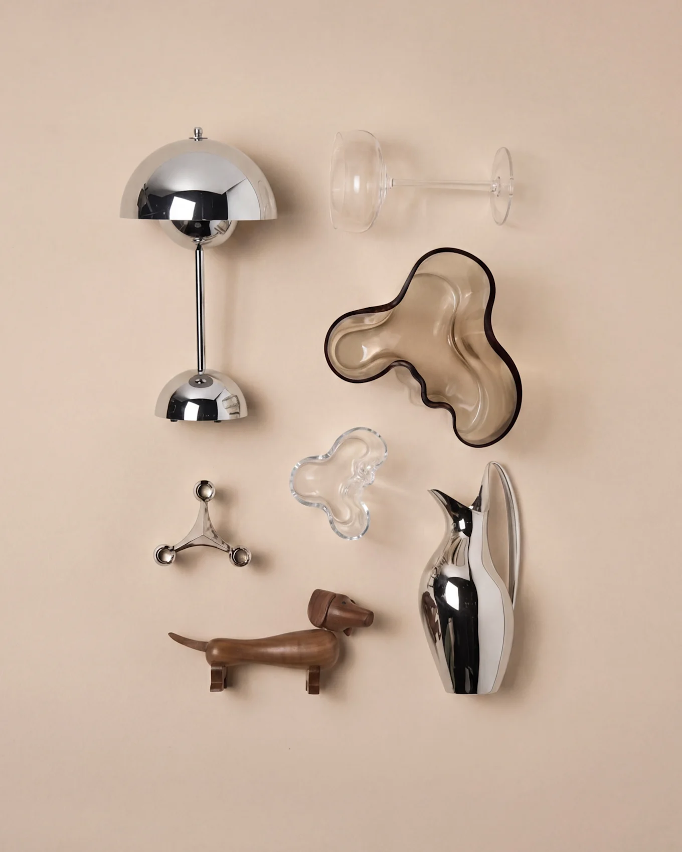 Modern home decor: chrome mushroom lamp, clear wine glass, brown wavy bowl, silver candle holder, wooden dachshund, silver pitcher.