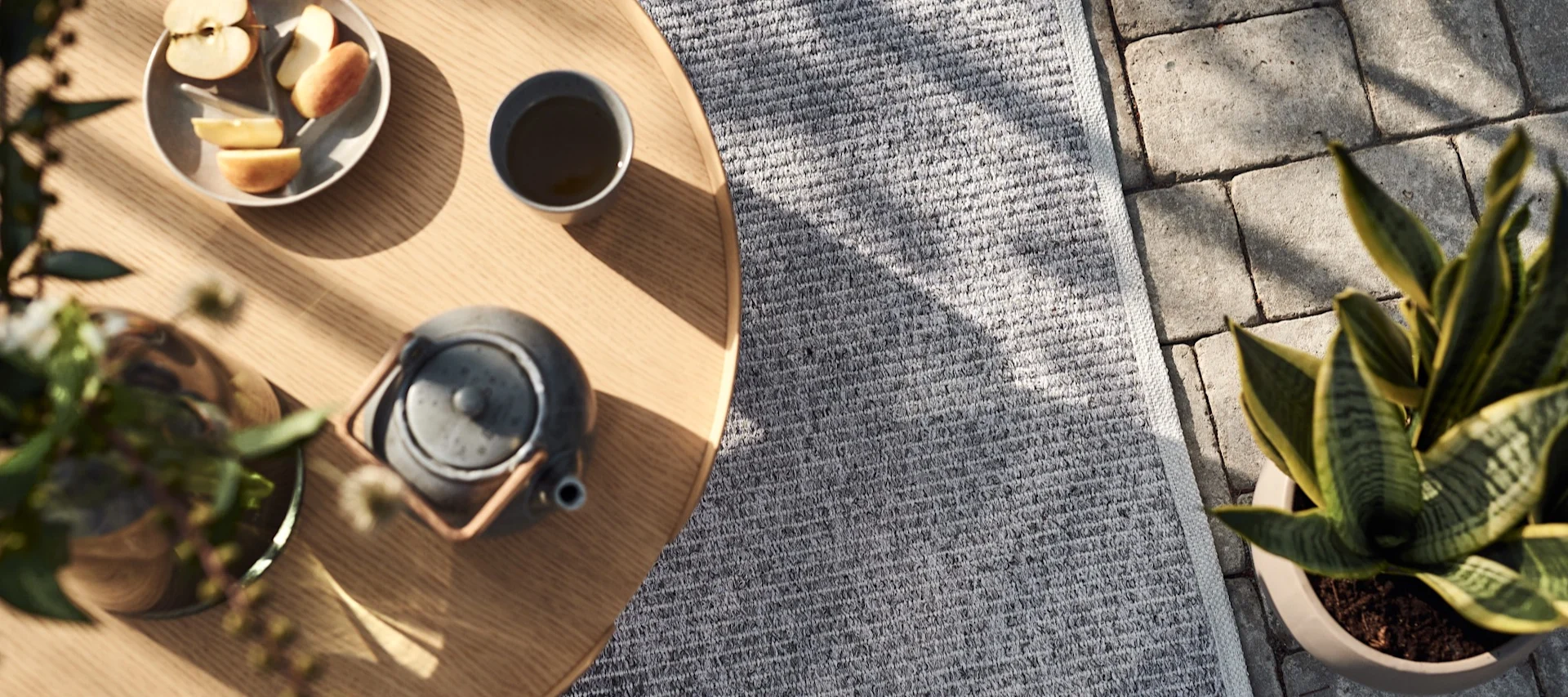 A wooden outdoor table with a plate of sliced apples, a cup of tea, and a teapot next to a grey rug and a snake plant.