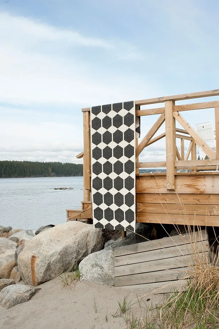 A black and white hexagon-patterned rug hangs over a wooden deck railing by a rocky, sandy shore.