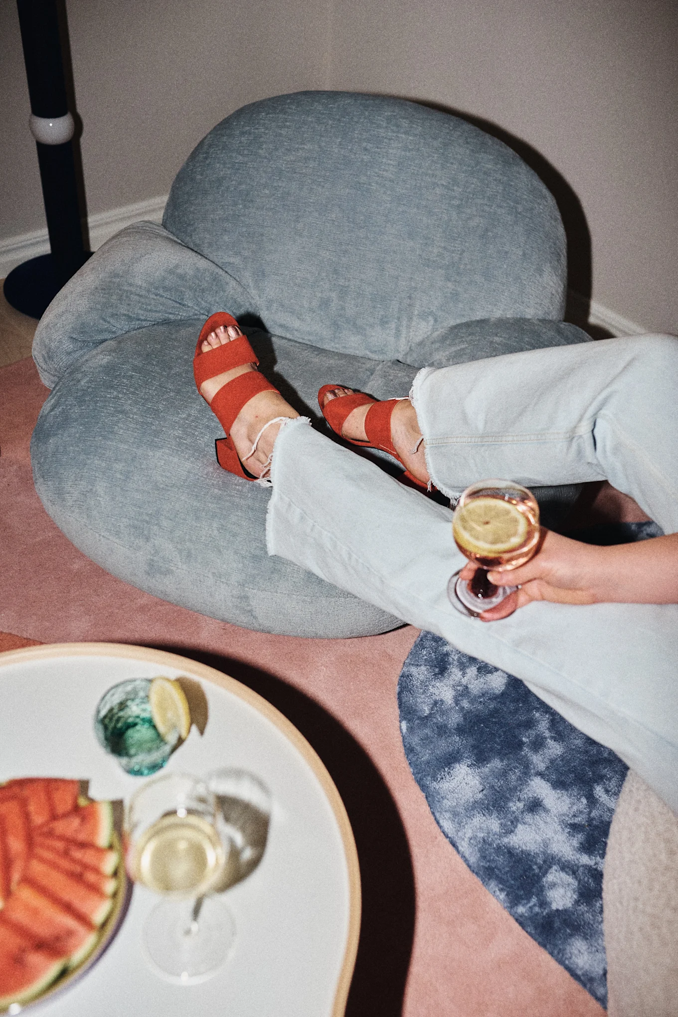 Person wearing light blue jeans and red sandals, relaxing on a blue chair with a drink and watermelon nearby.