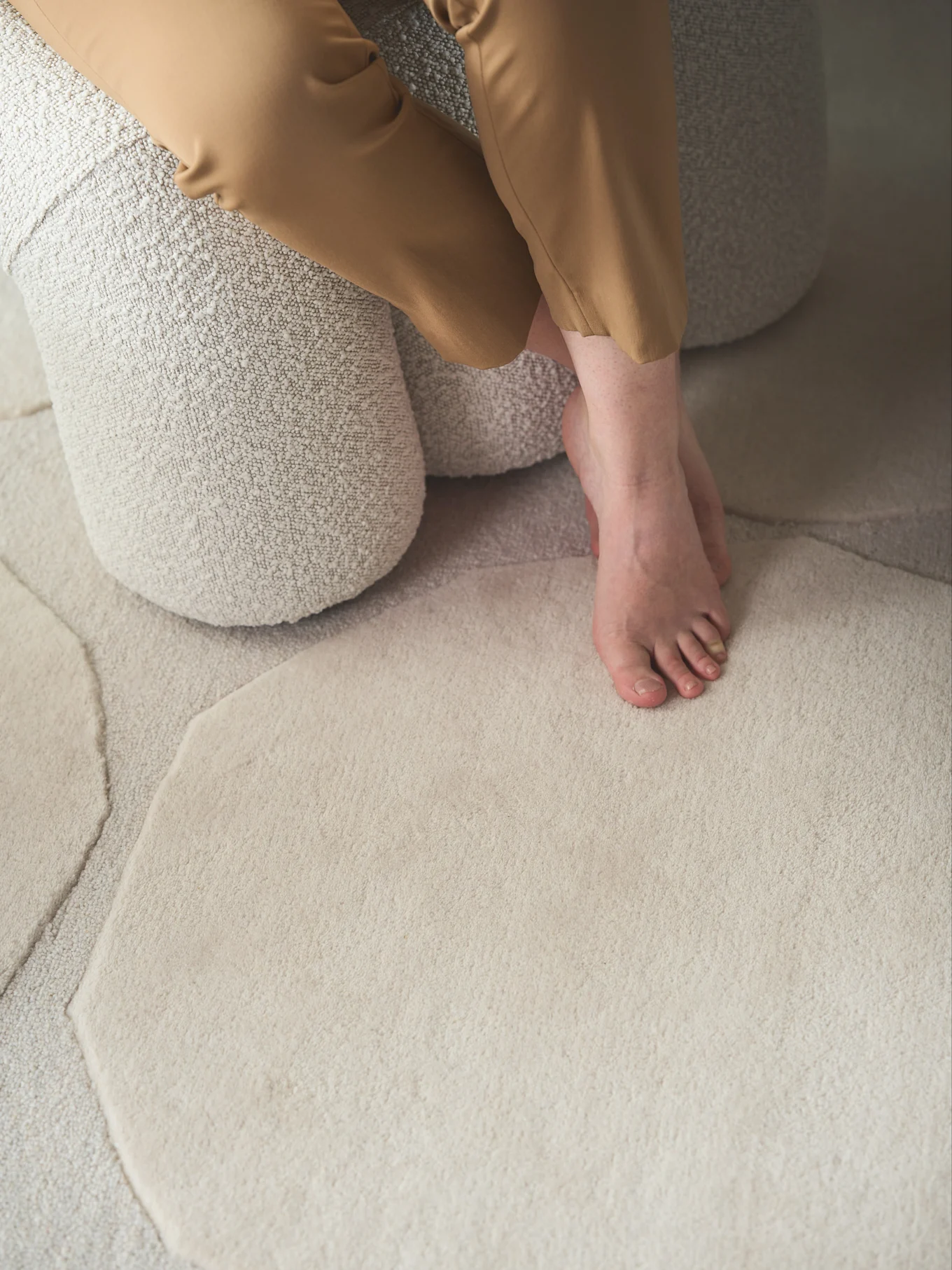 Bare feet and light brown trousers resting on a textured light beige rug with irregular shapes.