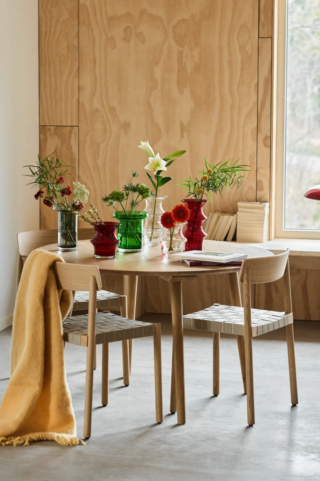A dining table in the middle of a plywood room with a collection of red, green, grey and clear vases. 
