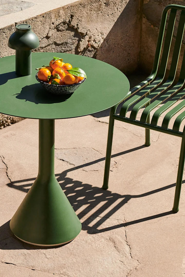 Green outdoor dining set with a round table, mushroom lamp, and bowl of tangerines on a sunlit patio.